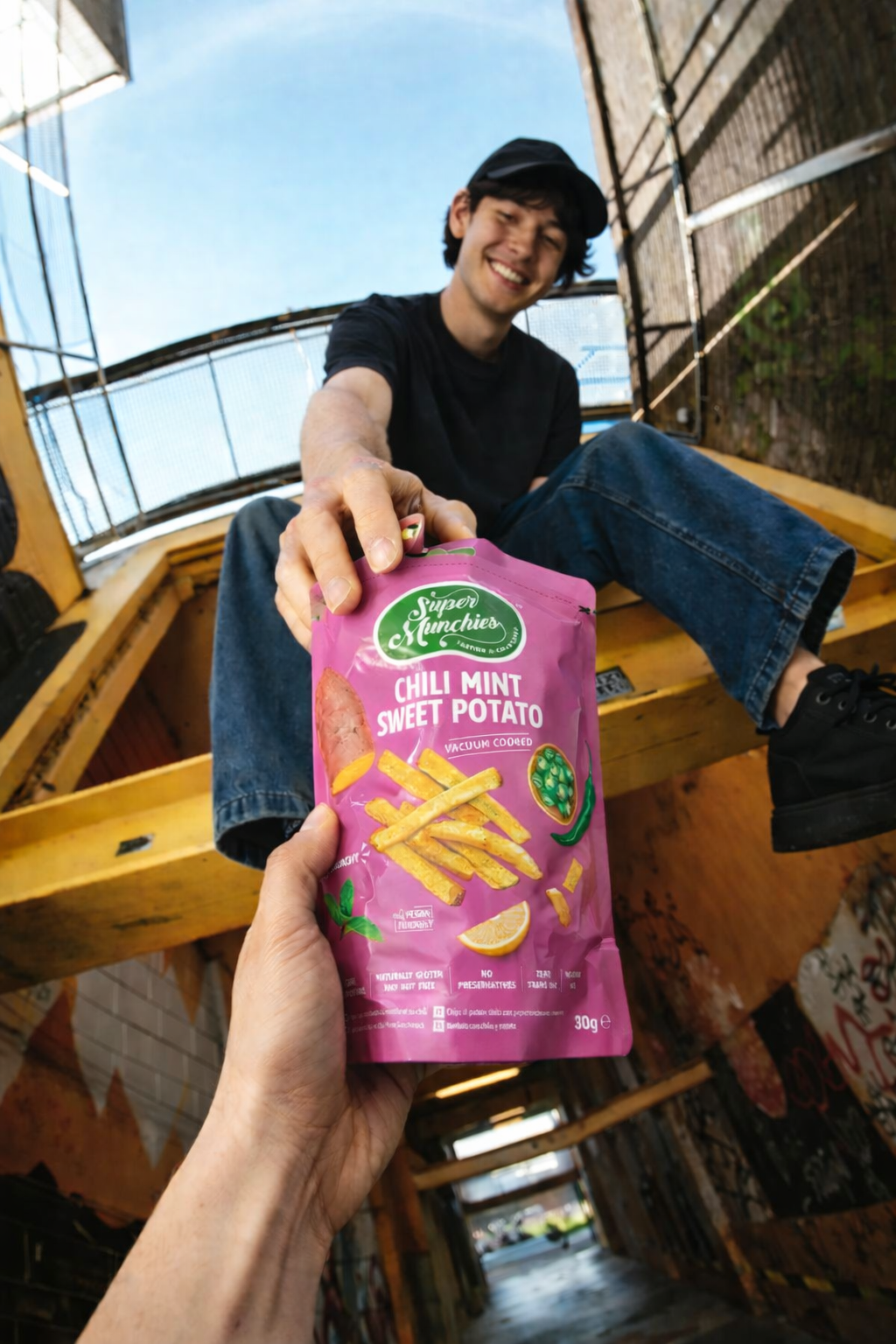 Mężczyzna siedzi na schodach i podaje opakowanie chipsów Chili Mint Sweet Potato Super Munchies, chrupiąca przekąska gotowana próżniowo