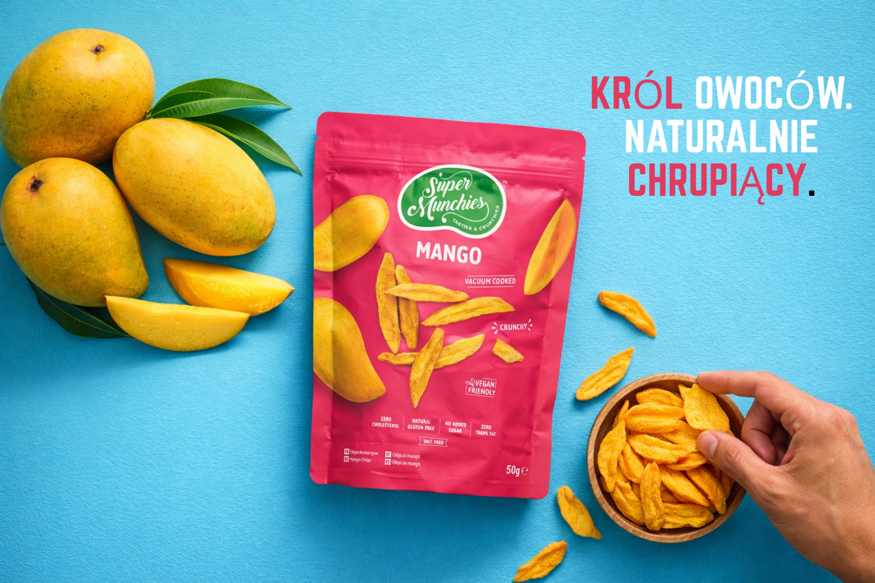 Opakowanie chipsów z mango Super Munchies obok świeżych mango i miseczki z chipsami na niebieskim tle z hasłem „Król owoców naturalnie chrupiący”
