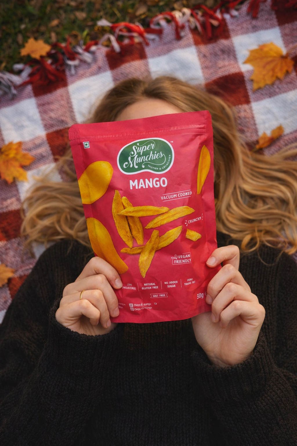 Kobieta leży na kocu i trzyma przed twarzą opakowanie chipsów z mango Super Munchies, naturalnie słodka i chrupiąca przekąska