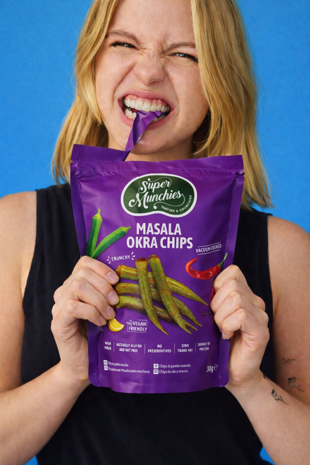 Kobieta otwiera opakowanie chipsów z okry Masala Super Munchies, chrupiąca przekąska wysokobłonnikowa i źródło białka na niebieskim tle