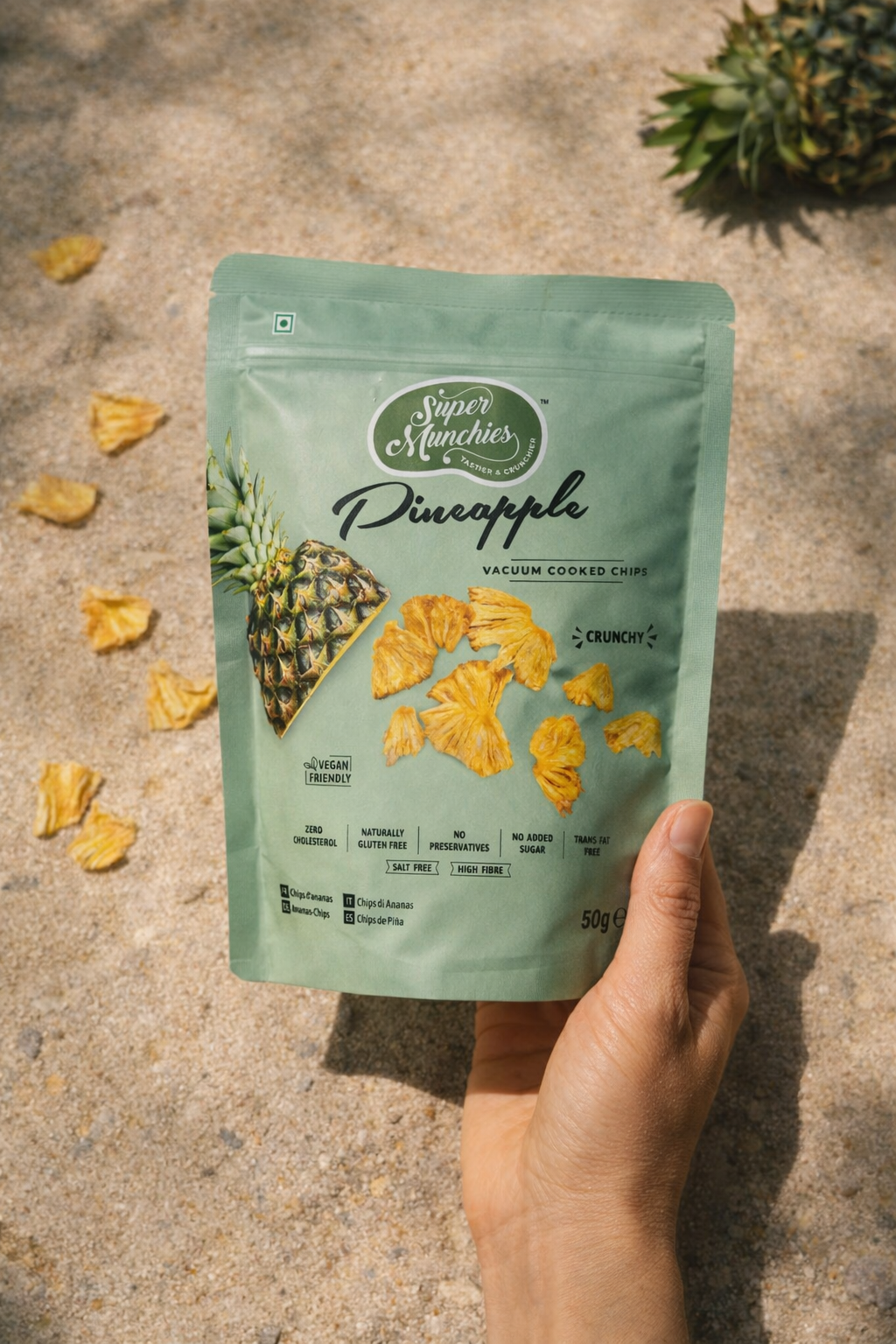 Dłoń trzyma opakowanie chipsów z ananasa Super Munchies na piasku, chrupiące chipsy gotowane próżniowo